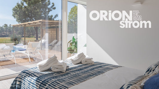 Φωτεινό υπνοδωμάτιο ξενοδοχείου με κρεβάτι στρωμένο ORION Strom, λευκές πετσέτες διπλωμένες και θέα σε ξύλινη βεράντα με ξαπλώστρες και πράσινο τοπίο.